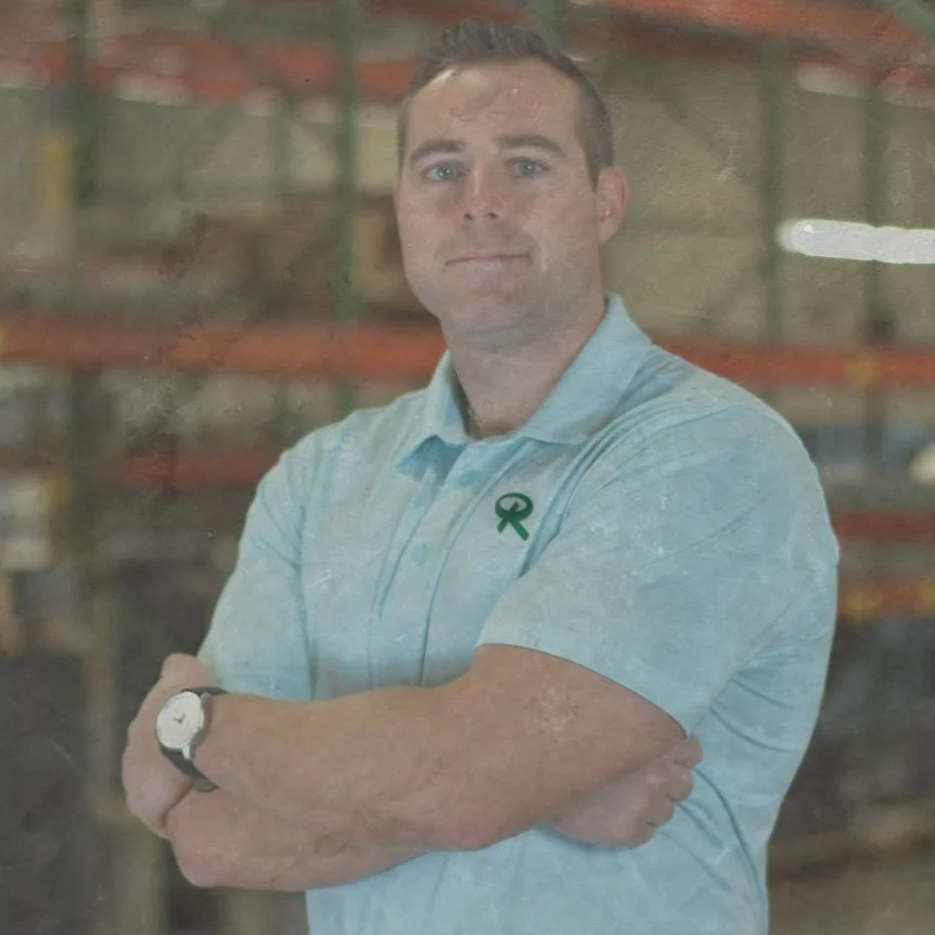 A person in a light blue polo shirt with a green ribbon emblem, standing with arms crossed.