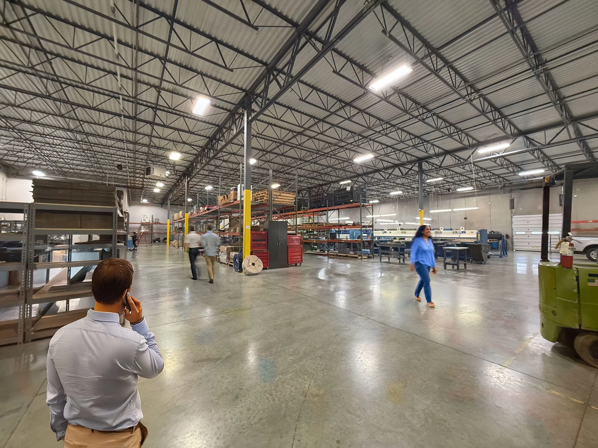 Large industrial warehouse with people walking, metal shelves, and machinery; man on phone in foreground.